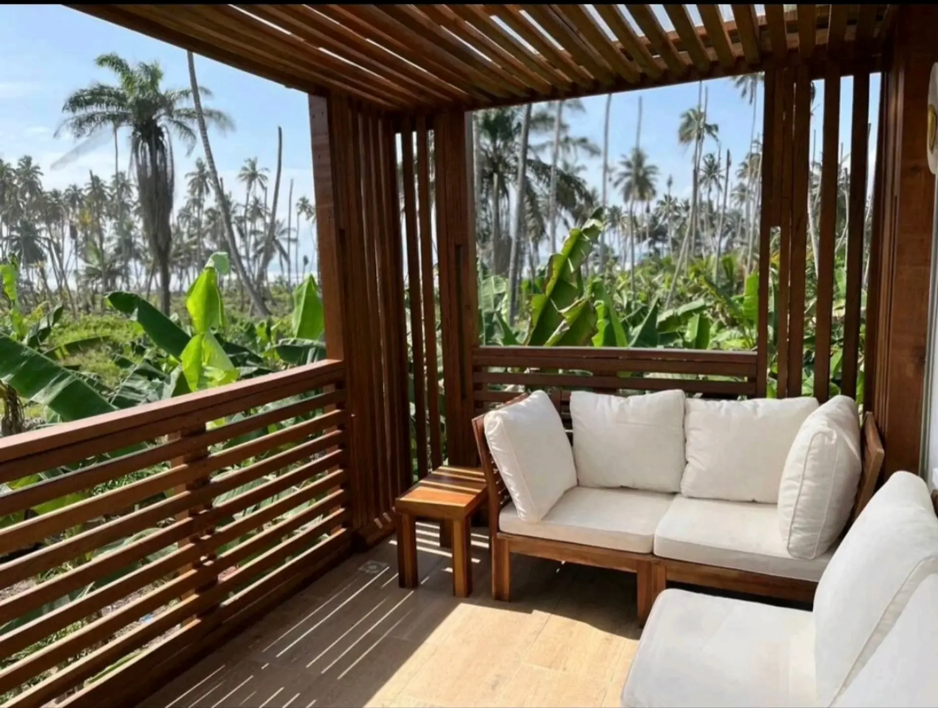 Covered wooden terrace with cushioned seating looking onto coconut palms